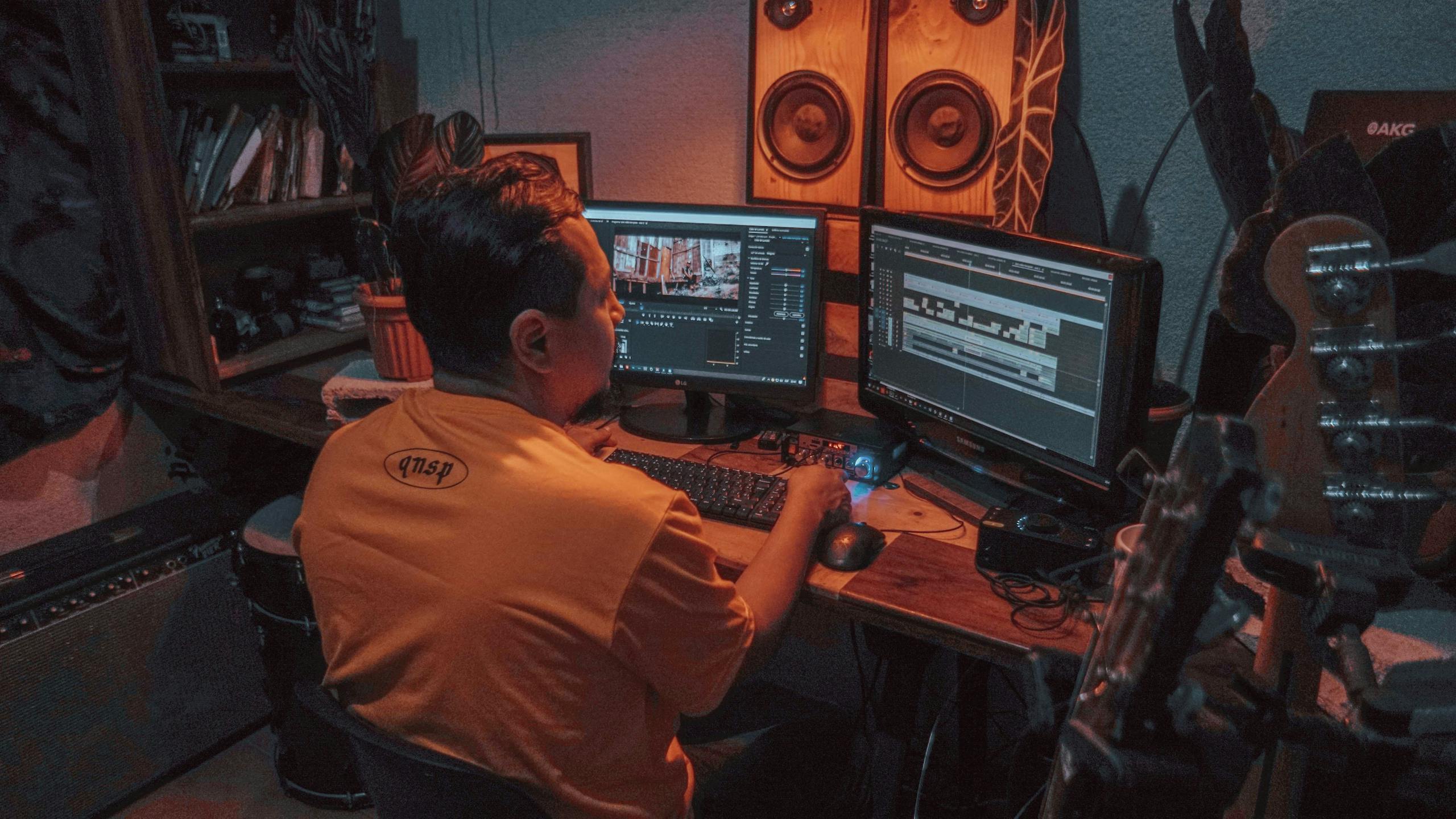 A person is editing video footage on dual monitors in a home studio with music equipment.